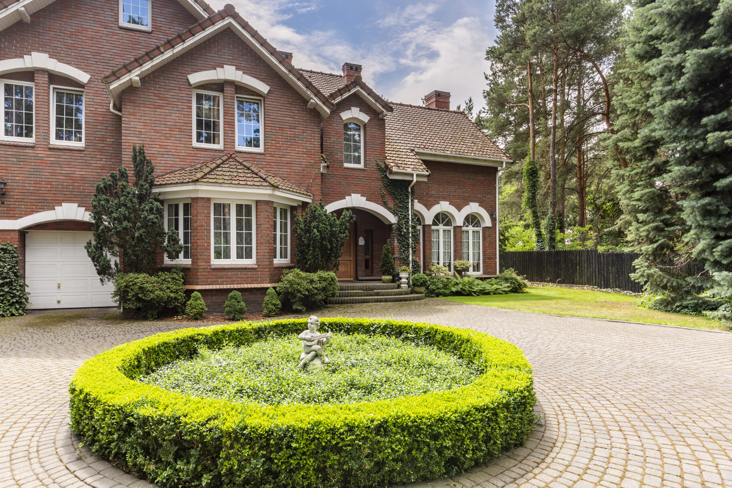 Round hedge and a small statue in the middle of a cobblestone driveway to an elegant, english style house with white windows and brick exterior